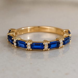 Puede incluir: Un anillo de banda dorada que presenta una serie de piedras azules de corte baguette alternadas con pares de pequeñas piedras redondas transparentes, dispuestas en un patrón horizontal a lo largo del metal para un diseño continuo.