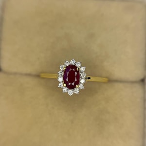 May include: A gold ring featuring a large oval ruby gemstone surrounded by a halo of small, round diamonds. The ruby is a deep red color, and the diamonds sparkle against the gold band. The ring is set in a classic, elegant style.