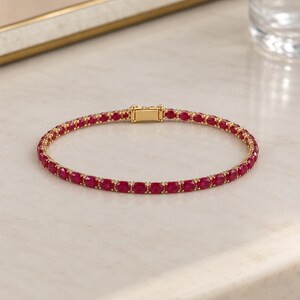 Könnte beinhalten: Ein goldenes Tennisarmband mit einer durchgehenden Reihe oval geschliffener, leuchtend roter Rubine in einer polierten Goldfassung. Das Armband verfügt über einen sicheren, rechteckigen Goldverschluss, der die beiden Enden des Schmuckstücks verbindet.