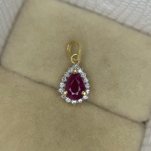 Könnte beinhalten: Ein goldener Anhänger mit einem tropfenförmigen, tiefroten Edelstein, umgeben von einem Kranz aus kleinen, klaren Steinen. Der Anhänger hat eine goldene Öse für eine Kette. Die Fassung ist aus Gold.