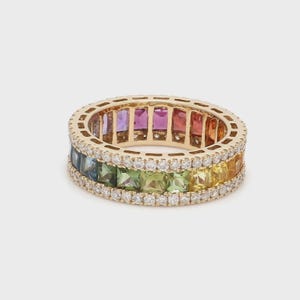 May include: A gold ring with a rainbow of gemstones. The ring features a band of colorful gemstones, including blue, green, yellow, and red, and is accented with a row of small, clear stones. The ring is set against a neutral background.