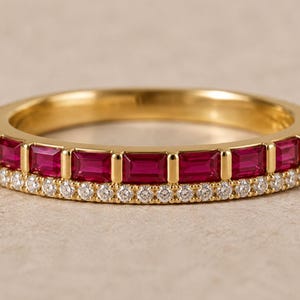 May include: A gold band ring featuring a row of rectangular baguette-cut red rubies set vertically, accented by a parallel row of small round white diamonds along the bottom edge of the band, creating a dual-textured metallic finish.