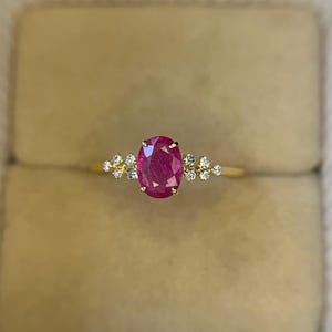 May include: A gold ring featuring a large, oval-cut ruby gemstone as the centerpiece. The ruby is a vibrant red color. Small, round, clear gemstones are clustered on either side of the ruby. The ring is set against a neutral background.