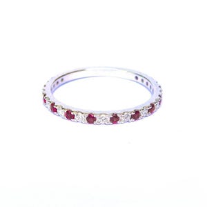 May include: A silver ring featuring a continuous row of alternating red and clear gemstones. The ring is set against a white backdrop, accentuating the sparkle of the stones. A delicate piece of jewellery.