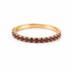 May include: A gold ring with a band of small, round, deep red gemstones. The ring is set against a white background, highlighting the gold and the red stones. The ring is a simple, elegant design.