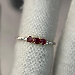 Op de afbeelding: Een delicate ring met drie levendige rode edelstenen gezet in een gouden band. De band is geaccentueerd met kleine, heldere stenen. De ring wordt gepresenteerd tegen een witte achtergrond, wat de glans en het ontwerp van het sieraad benadrukt.