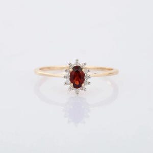 May include: A gold ring featuring a central oval-cut, deep red gemstone, surrounded by a halo of smaller, clear gemstones. The ring's band is a thin, polished gold. The design is elegant and classic, suitable for various occasions.