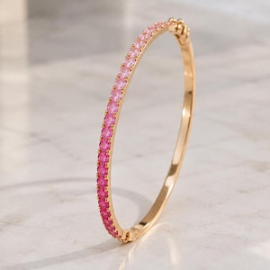 May include: A thin gold bangle bracelet featuring a continuous row of small, round, vibrant pink gemstones set along the outer edge. The bracelet includes a small, integrated hinged clasp mechanism at the top for opening and closing.