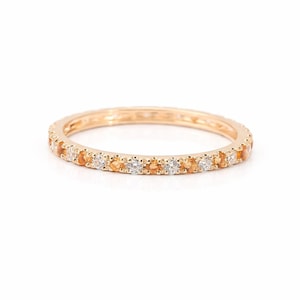 Könnte beinhalten: Ein goldfarbener Ring mit einem Band aus abwechselnd orangefarbenen und weißen Edelsteinen. Der Ring ist vor einem weißen Hintergrund platziert, der das Funkeln der Edelsteine hervorhebt. Ein filigranes Schmuckstück.