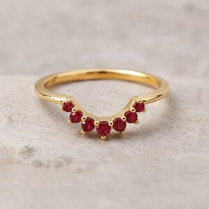 Könnte beinhalten: Ein goldener Ring mit einem geschwungenen, V-förmigen Mittelteil, der mit sechs kleinen, runden, leuchtend roten Rubinen besetzt ist. Der polierte Goldring hat eine glatte Oberfläche und die Steine sind in einem symmetrischen Bogen angeordnet.
