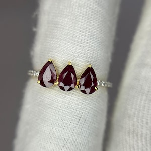 Könnte beinhalten: Ein goldener Ring mit drei tropfenförmigen, tiefroten Edelsteinen. Die Edelsteine sind in einer Reihe angeordnet, wobei kleinere, klare Steine das Band akzentuieren. Der Ring wird auf einem weißen Stoffhintergrund präsentiert.