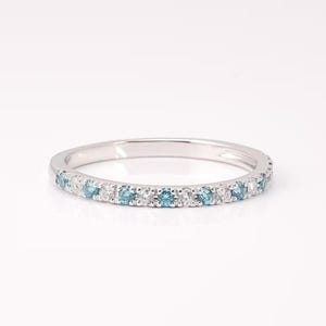 May include: A silver band ring with alternating blue and white gemstones. The ring is set with small, round-cut stones, creating a continuous row of sparkle. The ring is displayed on a white background, highlighting its delicate design.