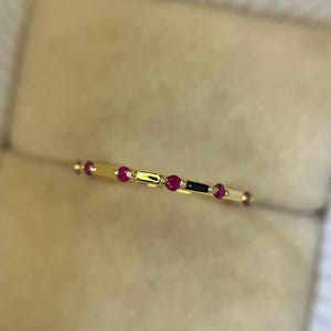 Può includere: Un delicato anello d'oro con piccole pietre preziose rotonde di rubino. L'anello ha una sottile fascia con segmenti d'oro e accenti di rubino alternati. L'anello è esposto su uno sfondo beige neutro.