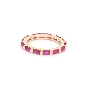 May include: A gold-colored ring with a continuous band of rectangular, bright pink gemstones. The ring is set against a white background, highlighting the vibrant color and design of the jewelry.