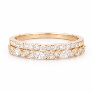 May include: Two gold rings stacked together. The top ring is lined with small, round, clear stones. The bottom ring features a mix of round and marquise-cut clear stones, creating a sparkling effect. The rings are set against a white background.