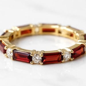 Op de afbeelding: Een gouden eeuwigheidsring met afwisselend baguette-geslepen dieprode granaatstenen en ronde, briljant-geslepen helderwitte diamanten in een doorlopend cirkelvormig patroon. De metalen band heeft een gepolijste afwerking en een constante breedte.