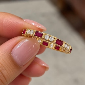 May include: A gold eternity band featuring an alternating pattern of rectangular baguette-cut red rubies and pairs of round brilliant-cut white diamonds set in a continuous row around the entire circumference of the metal ring band.