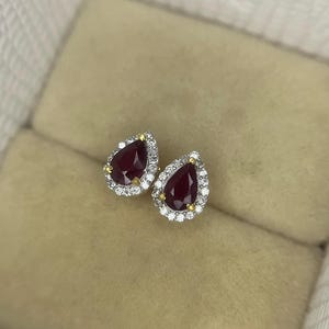 Puede incluir: Un par de pendientes en forma de lágrima. Cada pendiente presenta una piedra preciosa de color rojo intenso rodeada por un halo de pequeñas piedras transparentes. El engaste parece ser de oro y plata, creando un contraste de colores. Los pendientes se exhiben sobre una superficie beige.