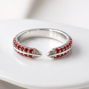 May include: A silver-toned open ring featuring two parallel rows of small, round, vibrant red stones set along the band. The ring has a bypass design with pointed ends that do not meet, showcasing a polished metallic finish and bright red accents.
