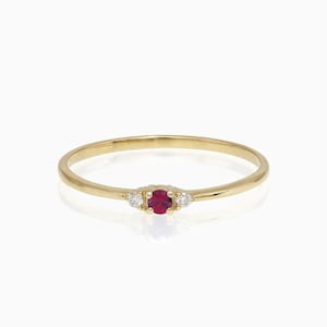 May include: A slender gold ring with a central oval ruby, set between two smaller, clear gemstones. The ring has a thin, polished gold band. The gemstones are arranged symmetrically, creating a classic design.