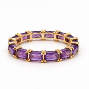 Op de afbeelding: Een gouden eternity-ring met een doorlopende rij rechthoekige, baguette-geslepen paarse amethist edelstenen. Elke edelsteen wordt op zijn plaats gehouden door kleine gouden pootjes, wat zorgt voor een uniform, cirkelvormig ontwerp.