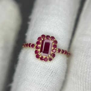 Puede incluir: Un anillo de oro con un gran rubí rectangular central, rodeado de rubíes más pequeños. La banda también está adornada con pequeños rubíes. El anillo se presenta sobre un fondo blanco, resaltando las vibrantes piedras rojas.