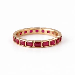 Könnte beinhalten: Ein goldener Eternity-Ring mit einer durchgehenden Reihe rechteckiger, im Treppenschliff facettierter, leuchtend roter Rubine, die in einen polierten Goldrahmen gefasst sind. Der Ring zeigt ein sich wiederholendes Muster aus Edelsteinen.