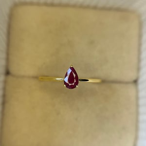 Könnte beinhalten: Ein goldener Ring mit einem tropfenförmigen, tiefroten Edelstein. Der Ring hat ein schmales Band, und der Edelstein ist das Hauptmerkmal, das in einem einfachen, eleganten Design gefasst ist. Der Ring wird in einer beigen Schmuckschatulle präsentiert.