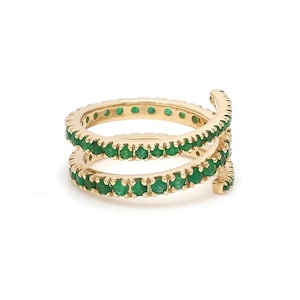 May include: A gold-coloured spiral ring featuring two bands of small, vibrant green gemstones. The ring is set against a white background, highlighting the contrast between the gold and the green stones. The design is elegant and modern.