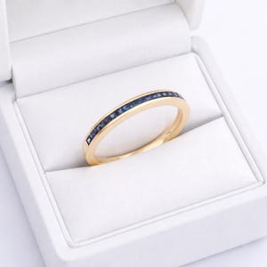 May include: A gold ring featuring a line of small, dark blue gemstones. The ring is presented in a white jewellery box with a textured lining. The gold band complements the blue gemstones, set against the white backdrop of the box.