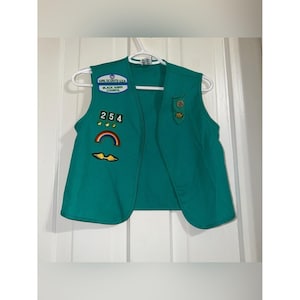 May include: A teal Girl Scouts USA vest featuring a Black Hawk Council patch, the number 254, three small gold star pins, a rainbow patch, and a yellow wing patch. The vest also includes two circular gold-toned pins on the left side.