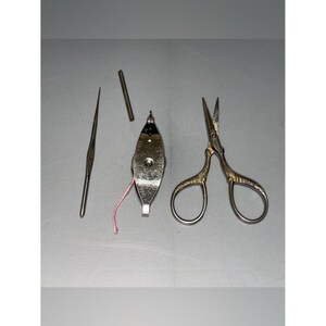 May include: A collection of vintage sewing tools, including a needle, a small metal tube, a thread holder with pink thread, and ornate gold-handled scissors. The tools are arranged on a gray surface.