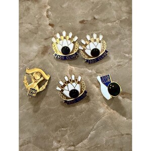 May include: Five gold-tone metal lapel pins featuring bowling themes. Four pins display white bowling pins and black bowling balls with blue banners reading 500 CLUB. One pin shows a gold figure with a banner reading 500 CLUB and a laurel wreath.