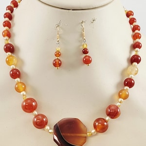 May include: A necklace and earring set featuring a variety of red, orange, and yellow beads. The necklace has a large, octagonal, brown pendant. The earrings have three beads each, with gold-colored hooks. The jewelry is displayed on a white mannequin.
