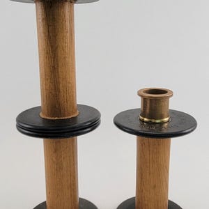 May include: Two wooden candle holders with brass tops and black bases. The candle holders have a classic design, with a tall, slender wooden body and a circular base. The brass tops are designed to hold candles. The candle holders are of different heights.