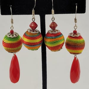 May include: Two pairs of dangle earrings featuring spherical beads wrapped in multicolored thread in shades of red, yellow, green, and blue. Each earring includes silver-toned metal filigree caps, a small red bead, and a faceted red teardrop pendant.