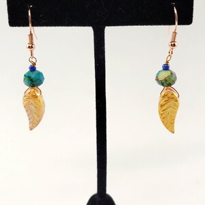 May include: A pair of dangle earrings featuring copper-toned hooks, small blue beads, faceted teal beads with dark veining, and gold-colored leaf-shaped charms. The earrings are suspended from a black T-shaped display stand.
