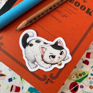 May include: A sticker featuring a cartoon cat with white fur, black spots, and a black and white head. The cat wears a red collar with a gold bell. The sticker is on an orange notebook with a pencil and blue pen.