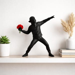 Peut inclure: Sculpture noire représentant une figure cagoulée et masquée, lançant un bouquet de fleurs rouges. La figure est en mouvement, un bras tendu. La sculpture est sur une étagère blanche avec une plante succulente et un vase.