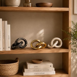 Peut inclure: Trois sculptures décoratives en forme de maillons de chaîne aux finitions noire, dorée et crème sont disposées sur une étagère en bois. Le maillon noir a une surface texturée, le doré est poli et brillant, et le crème a une finition mate lisse.