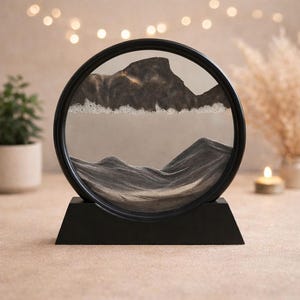Könnte beinhalten: Ein schwarzes, kreisförmiges Sandkunstwerk mit einem Berglandschaftsdesign. Der Sand ist in Grau- und Beigetönen gehalten und erzeugt einen dynamischen visuellen Effekt im Glas. Das Stück steht auf einem schwarzen Sockel und ist ein dekoratives Wohnaccessoire.