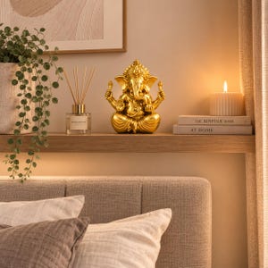 Peut inclure: Une figurine dorée métallique représentant la divinité Ganesh en position assise avec quatre bras, placée sur une étagère en bois à côté d'un diffuseur de parfum en verre et de deux livres empilés portant le texte The Kinfolk Home et At Home.