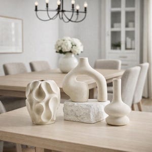 Peut inclure: Trois vases en céramique blanc cassé de formes et tailles variées sont exposés sur une table en bois clair. L'un a une forme bulbeuse, un autre une forme d'arche et le troisième une forme de bouteille élancée. Décor de salle à manger.