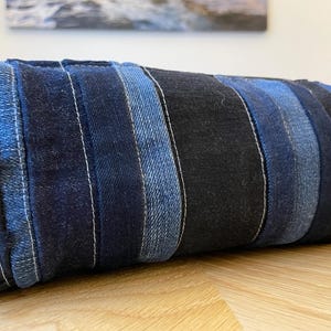 May include: A rectangular denim bag with vertical stripes of varying shades of blue. The bag is made from repurposed denim and stitched with white thread. The bag is resting on a wooden surface.