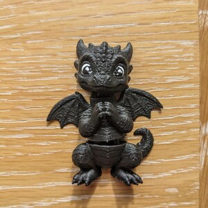 May include: A small, black, 3D-printed dragon figurine with textured scales, pointed ears, and small wings. The dragon has large, expressive eyes with white highlights, a rounded snout, and a curled tail. Its front limbs are held together near its chest.