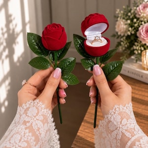 May include: Two red velvet rose-shaped ring boxes with green stems and leaves. One box is closed, while the other is open, revealing a gold ring with a central stone set inside the white interior of the rose-shaped container.