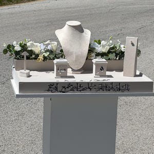 May include: A white jewelry display set featuring a necklace bust, two square ring boxes with cut-out Arabic calligraphy, a tall earring stand, and a small T-bar stand. The base is decorated with white calligraphic text and artificial white flowers.