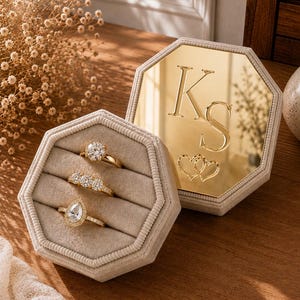 Puede incluir: Un joyero hexagonal de terciopelo beige con tres ranuras que contienen anillos dorados con piedras transparentes. La tapa es un espejo dorado con las letras KS grabadas y dos corazones entrelazados bajo las iniciales.