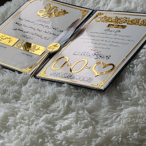 May include: Two wedding invitations with gold accents and Arabic script. The invitations are open, revealing ornate text and decorative elements. A pen with a feather is placed between the invitations. The background is a fluffy white surface.
