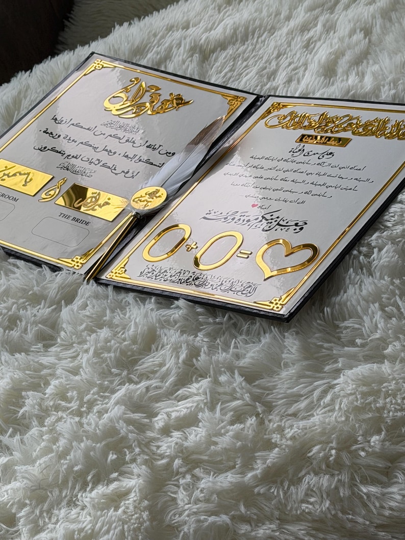 May include: Two wedding invitations with gold accents and Arabic script. The invitations are open, revealing ornate text and decorative elements. A pen with a feather is placed between the invitations. The background is a fluffy white surface.
